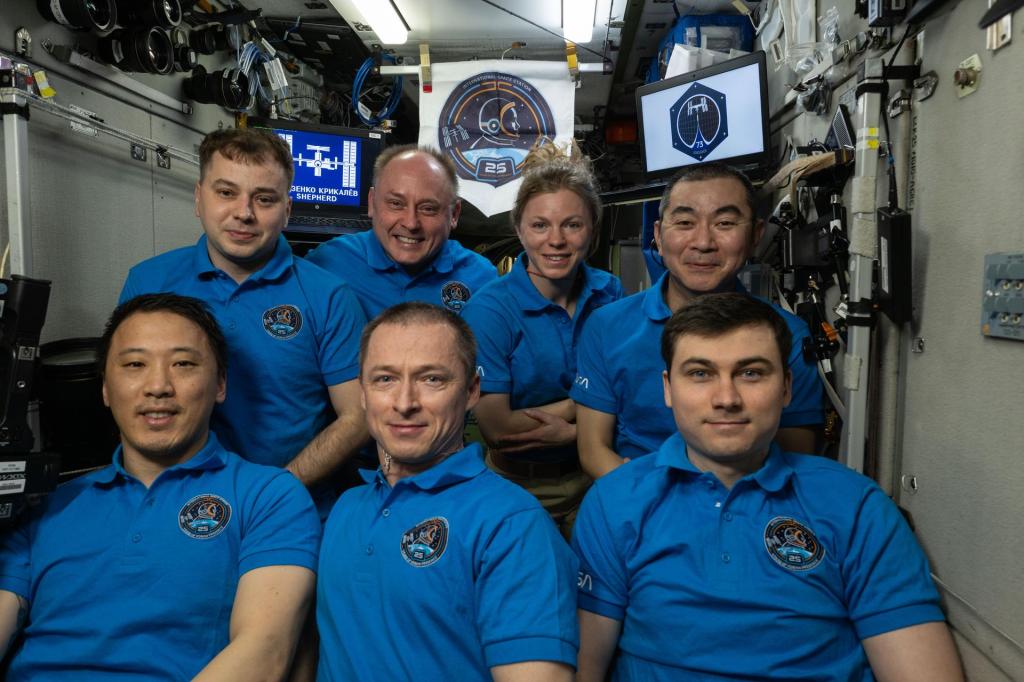 The seven-member Expedition 73 crew poses for a portrait on the International Space Station with two display screens behind them and a penant flag of the 25th anniversary logo identifier. The crew is wearing matching 25th anniversary polo shirts.