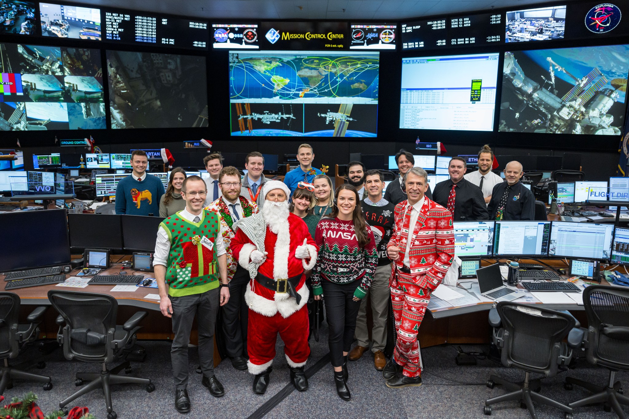 A group of people dressed in Christmas outfits pose for a photo in front of mission control.