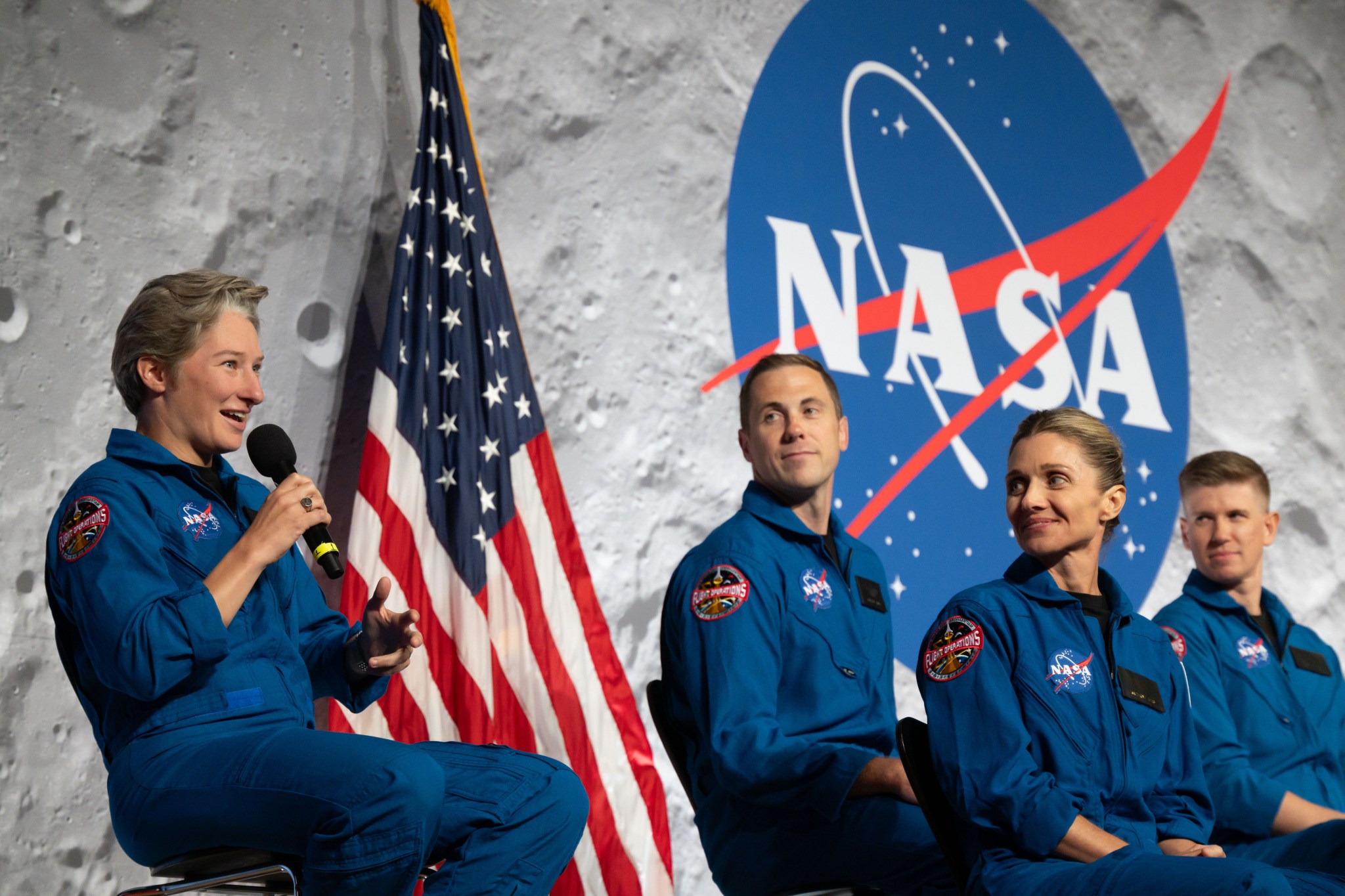 A woman speaks during a ceremony. Three people are beside her. The U.S. flag (left) and NASA meatball insignia are behind them.