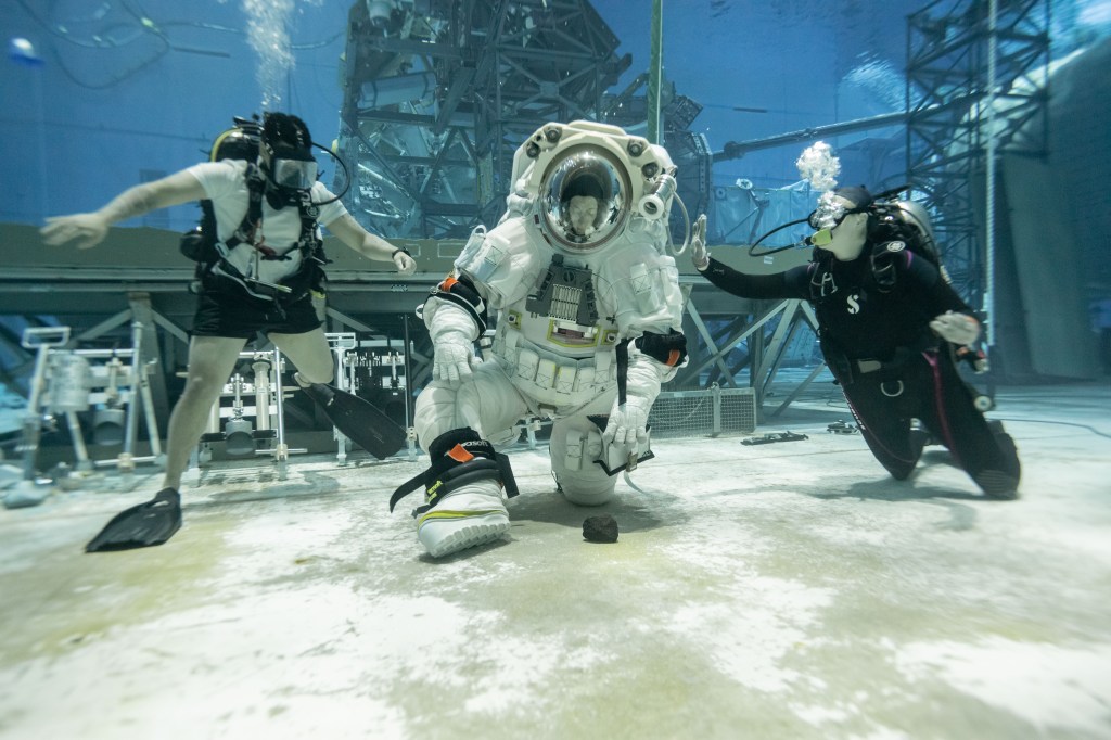 NASA astronaut Loral O'Hara kneels down to to pick up a dark lunar rock at the bottom of a pool during testing. O'Hara is wearing a white spacesuit while two scuba divers are on each side of her.