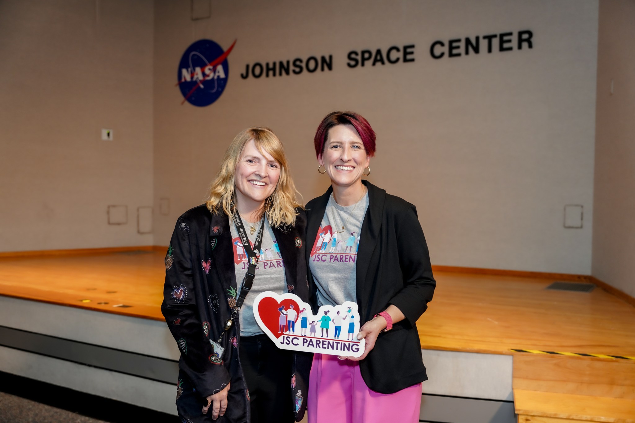 Two women hold a sign reading JSC Parenting while standing in front of a wooden stage.