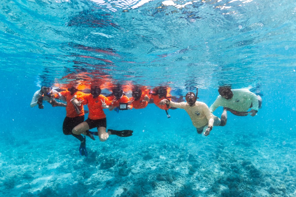 In this underwater shot, a group of students in bright orange long sleeves and flippers pose for the camera just underwater, next to a man in a cream long-sleeve and another man in a teal long sleeve.