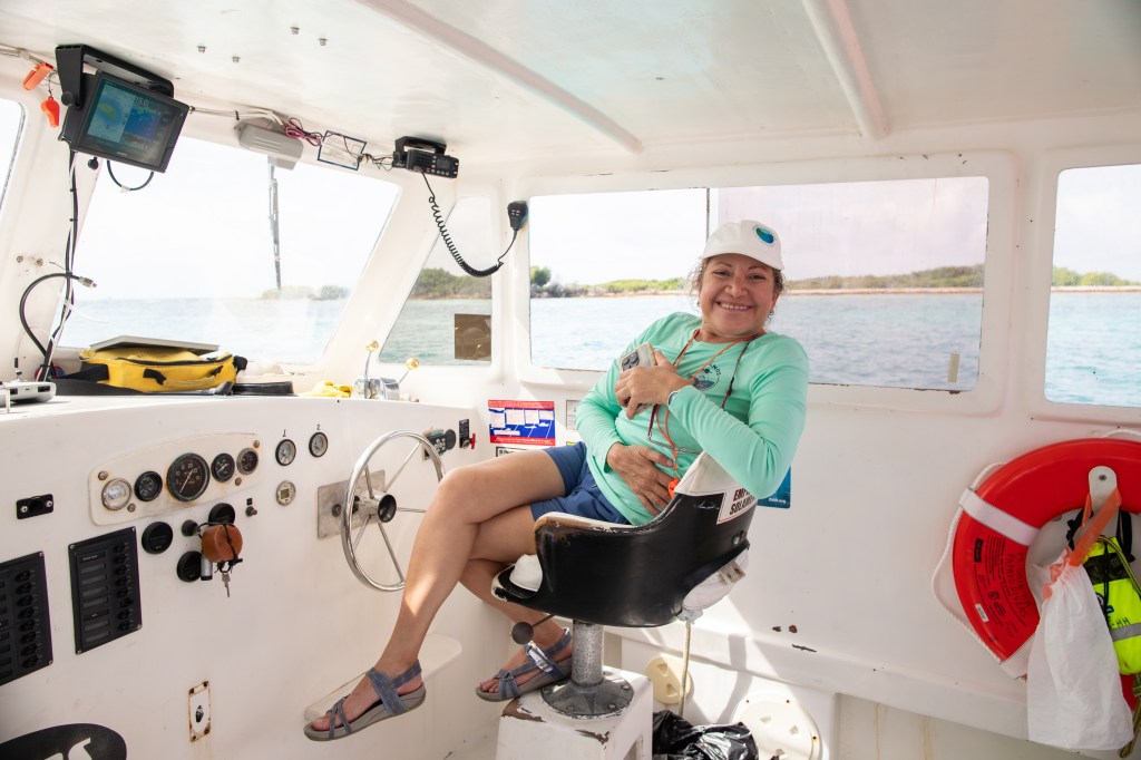 A woman in a teal long sleeve shirt, jean shorts, a white baseball cap, and sandals smiles at the camera from the captain's chair of a small boat. Behind her is the white interior of the boat, with a bit of water and land visible out the windows.