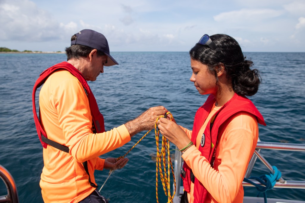 A man on the left hands rope to a young female intern on the right. Both are wearing bright orange long sleeve shirts and red life vests. In the background is blue ocean water.