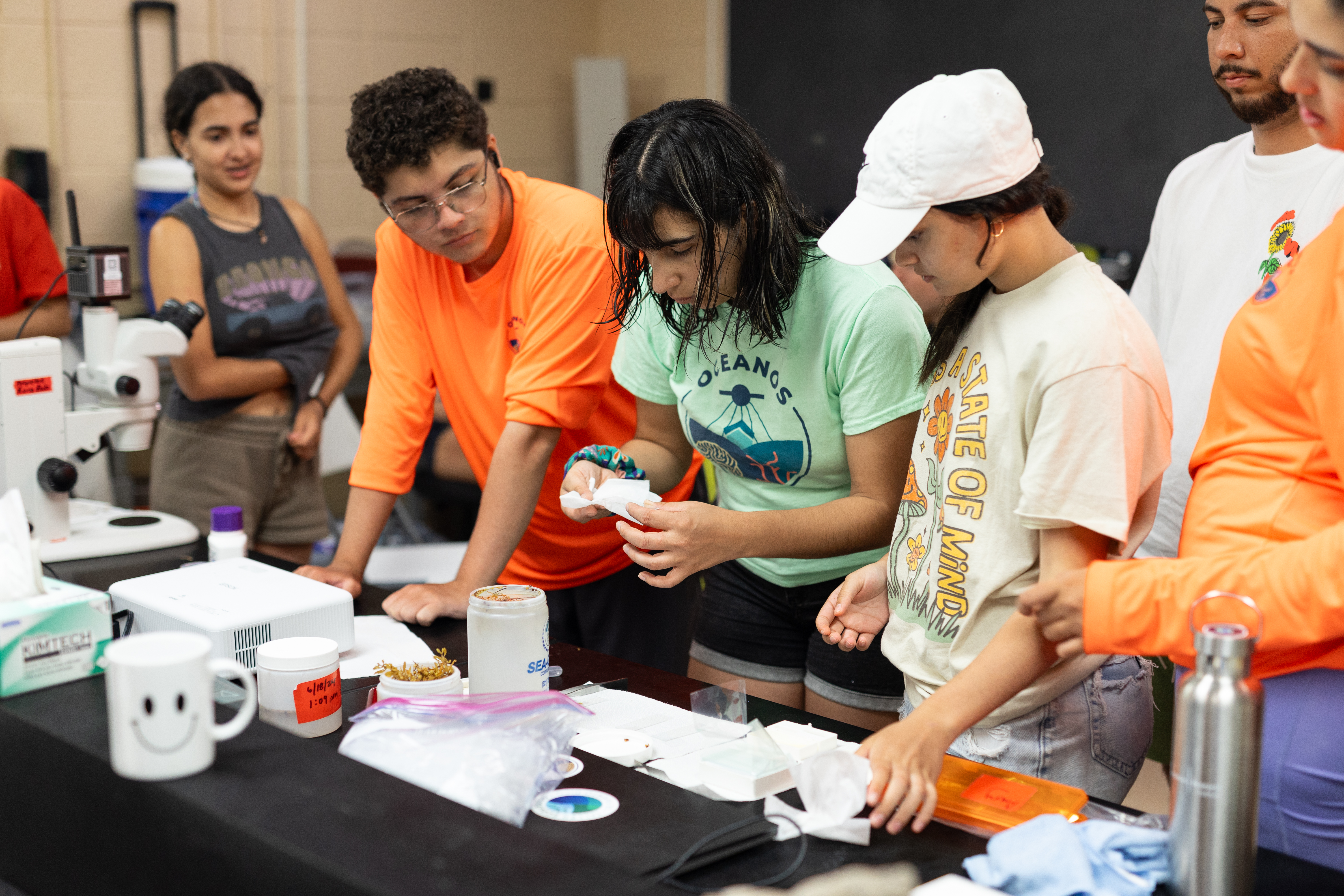 A group of students gather around a woman in a teal t-shirt with shoulder-length black hair as she bends over a lab table, holding a folded white cloth. On the lab table is an assortment of scientific lab materials and water bottles.