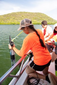 A girl in a neon orange long sleeve shirt, black shorts, a white baseball cap, with a long brown braid down her back, leans over the edge of a boat with a cylindrical instrument. In the background is green water and a mound of land covered in green bushes.
