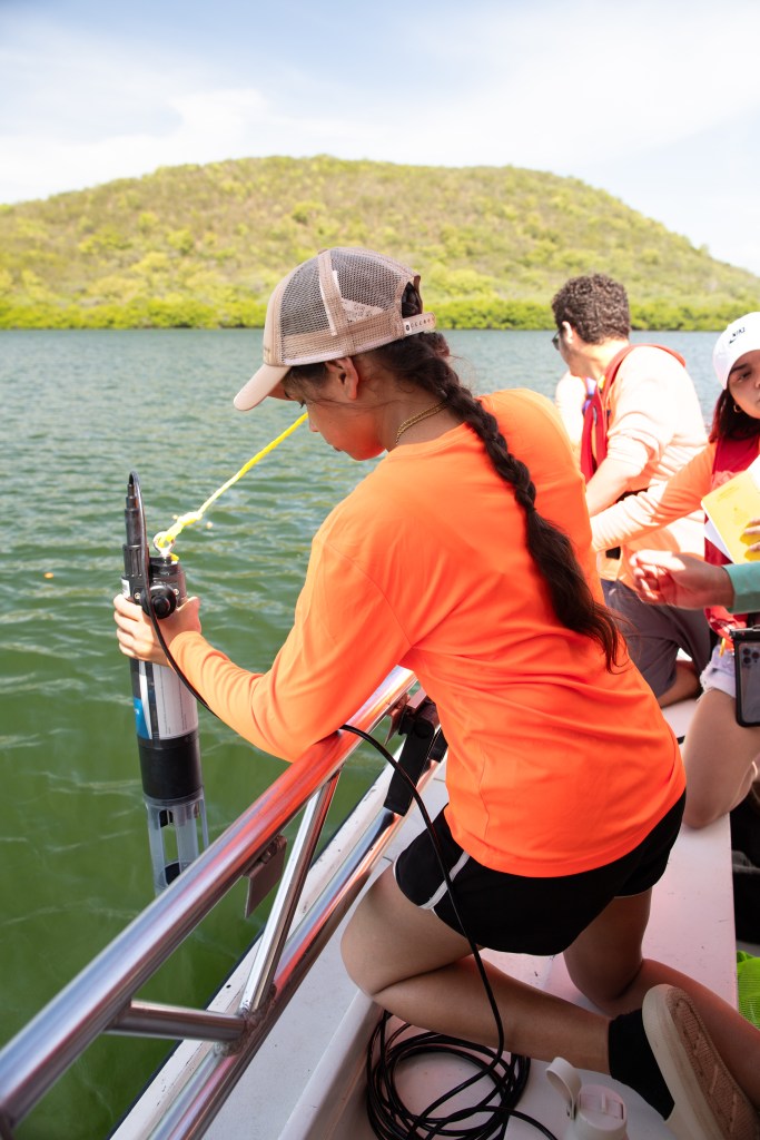 A girl in a neon orange long sleeve shirt, black shorts, a white baseball cap, with a long brown braid down her back, leans over the edge of a boat with a cylindrical instrument. In the background is green water and a mound of land covered in green bushes.