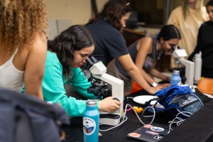 A woman in a bright teal long-sleeve shirt adjusts the side nob as she looks into a microscope. Around her are students gathered around lab tables, with one other microscope visible.