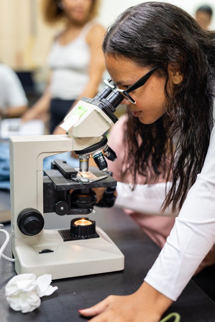 A woman with long dark hair, a white long sleeve shirt, and glasses looks through a microscope.