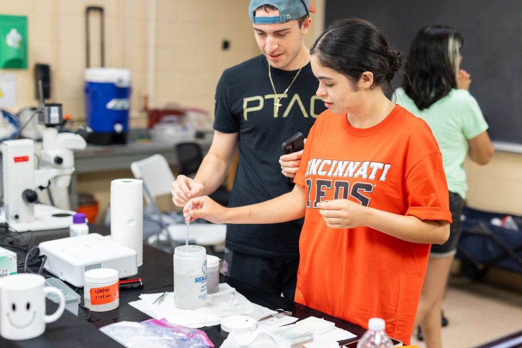 A man in a black t-shirt and backwards blue baseball cap stands next to a woman in an orange t-shirt that says Cincinnati Reds on the front. The woman is using a dropper to gather a sample from a white cylindrical vessel full of liquid. Various other scientific lab equipment is scattered on the black table in front of them.