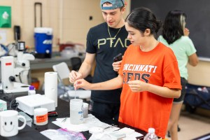 A man in a black t-shirt and backwards blue baseball cap stands next to a woman in an orange t-shirt that says Cincinnati Reds on the front. The woman is using a dropper to gather a sample from a white cylindrical vessel full of liquid. Various other scientific lab equipment is scattered on the black table in front of them.