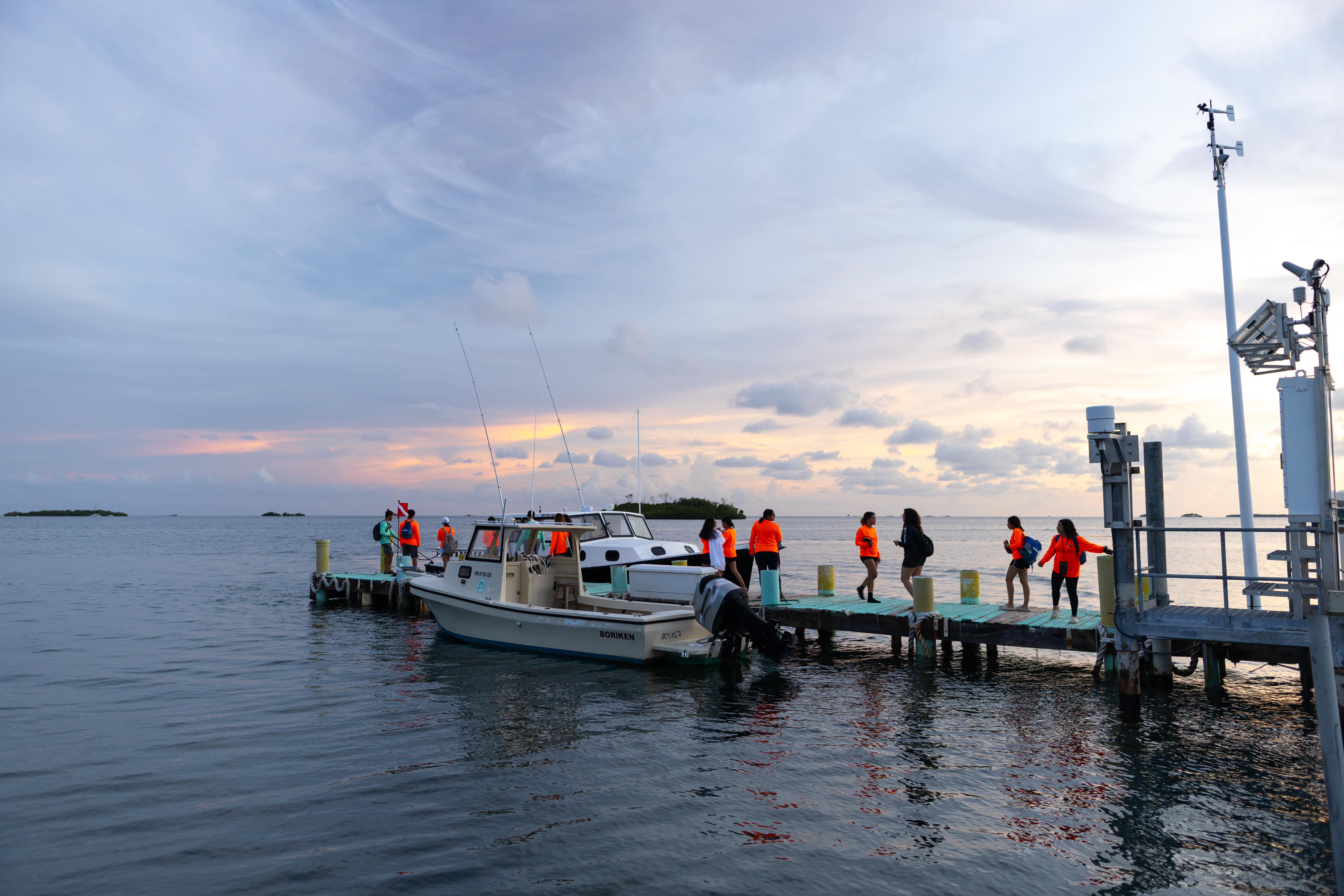 A pier stretches into the frame from the right, with a small white boat docked at the end. Students in neon orange long-sleeve shirts walk along the dock.