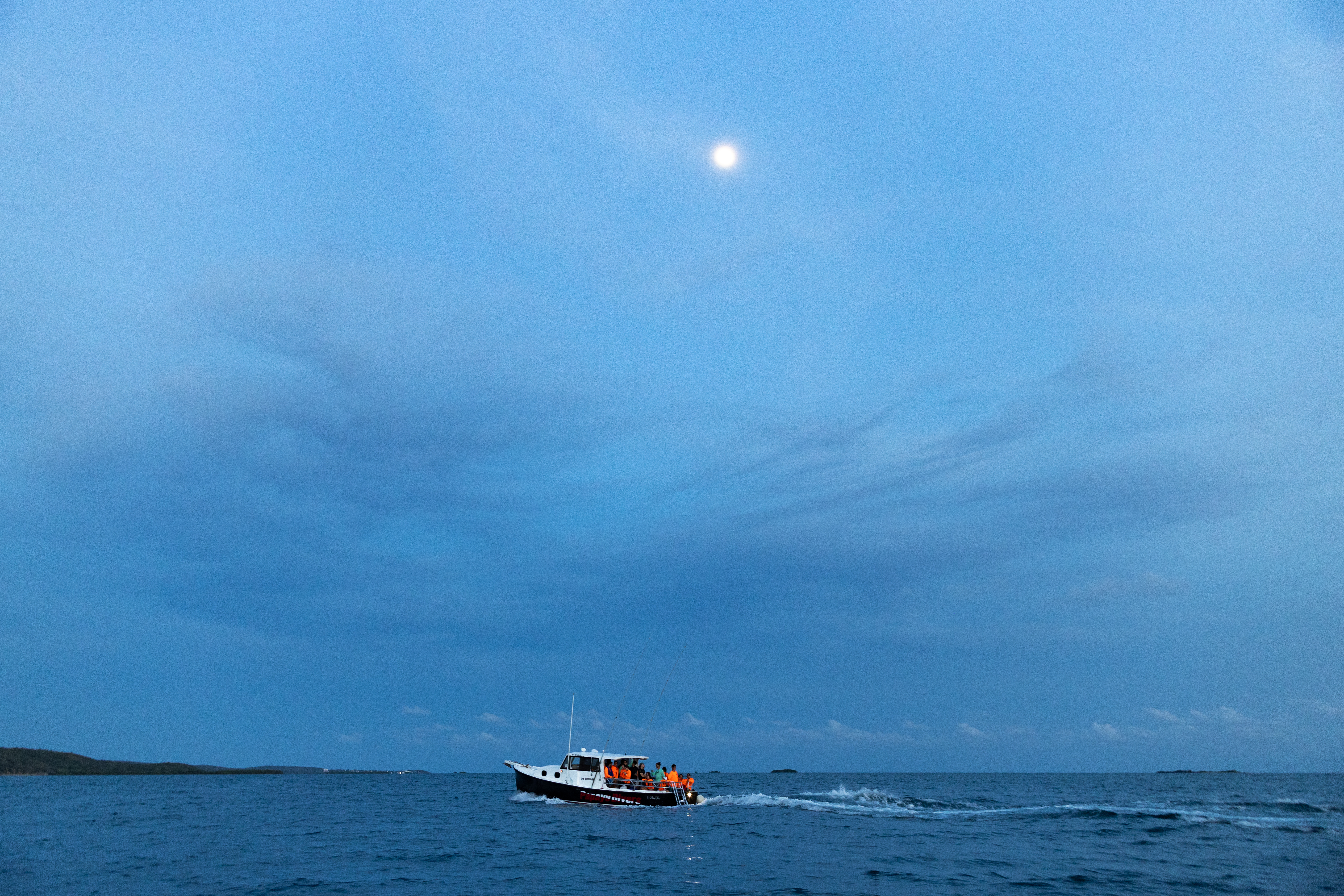 A distant blue-hour shot of a boat flying along the top of the water. Most of the photo is the blue sky with a few gray clouds, and a small moon glowing in the center top.