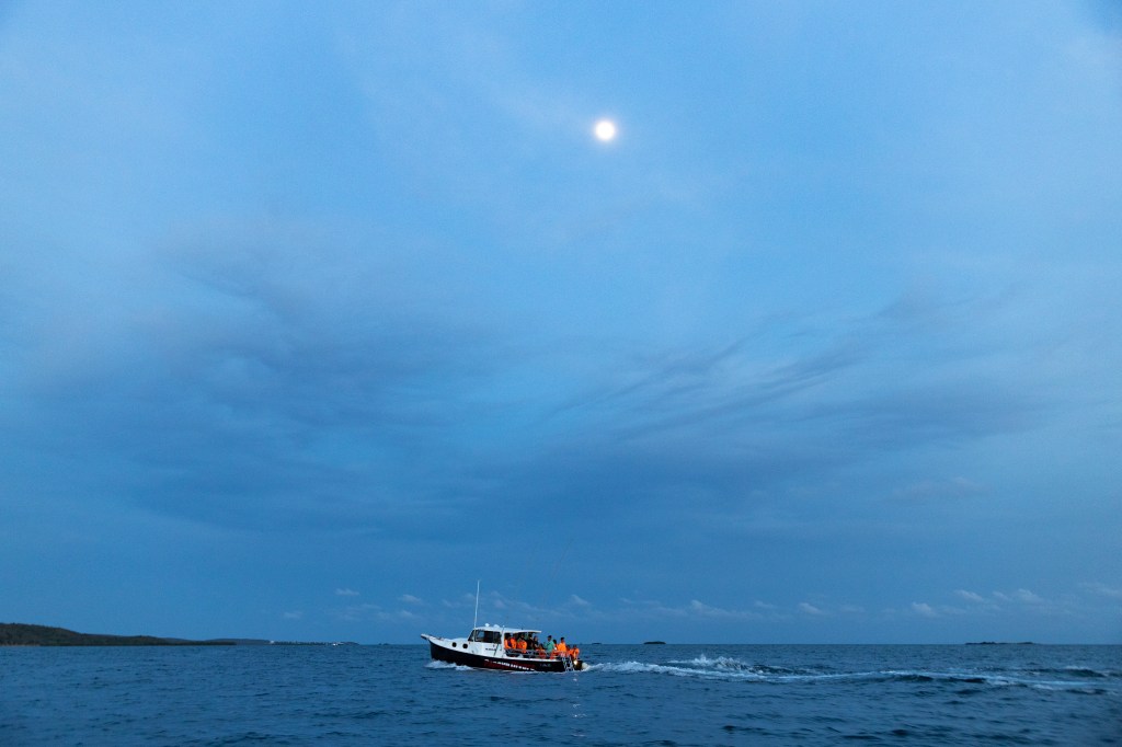 A distant blue-hour shot of a boat flying along the top of the water. Most of the photo is the blue sky with a few gray clouds, and a small moon glowing in the center top.