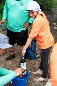 A student in a neon orange long sleeve shirt, gray leggings, and white head covering holds a cylindrical instrument into a bright blue bucket. She stands barefoot on a sandy beach.