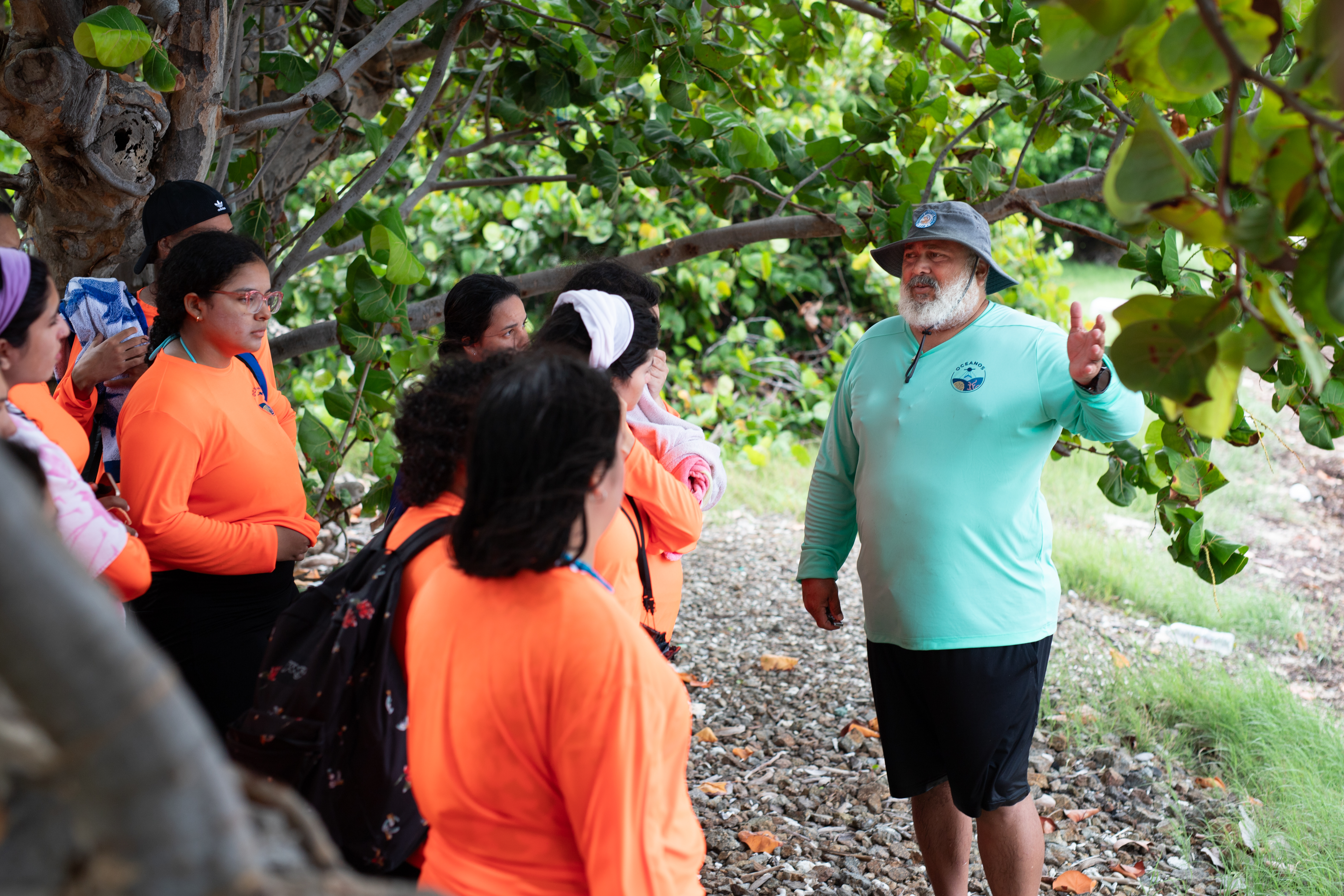 A man in a teal long sleeve shirt, black shorts, gray bucket hat, and long white beard gestures as he speaks to a group of students in orange shirts. The group stands on rocky ground, under a canopy of green leaves.