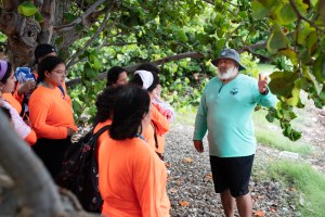 A man in a teal long sleeve shirt, black shorts, gray bucket hat, and long white beard gestures as he speaks to a group of students in orange shirts. The group stands on rocky ground, under a canopy of green leaves.