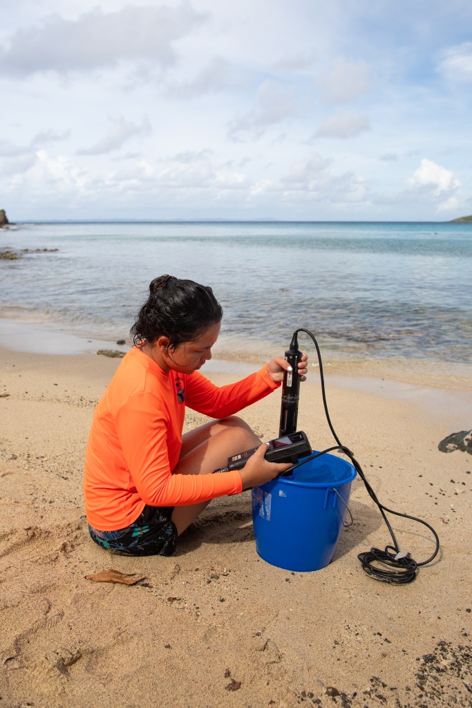 A girl in an orange long sleeve shirt with long dark hair in a ponytail sits on a beach, holding a black cylinder over a bright blue bucket. The ocean is visible in the background.