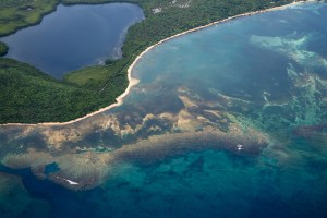 A photo from an aircraft looks down at the variegated blues, teals, tans, and browns of the ocean as it meets a stretch of dark green forested coastline. Inland a bit is a large dark blue lake.