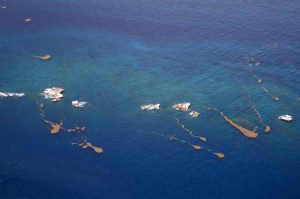 A photo from an aircraft looks down at the blended shades of blue and teal of the ocean, with a few spots of white and brown.