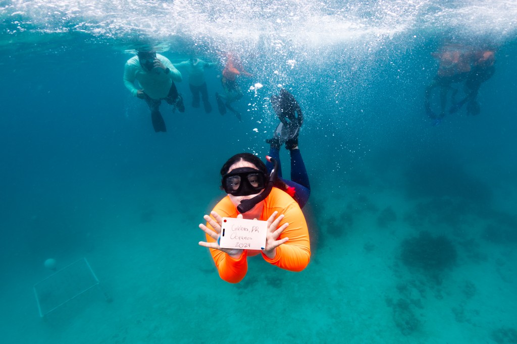 In this underwater image, a student in a neon orange long sleeve shirt, black leggings, fins, and a snorkel swims towards the camera, holding a small handwritten sign towards the camera that says 'Culebra, PR OCEANOS 2024.' The water all around her is bright blue, with the shadowy shapes of other snorkelers in the background.