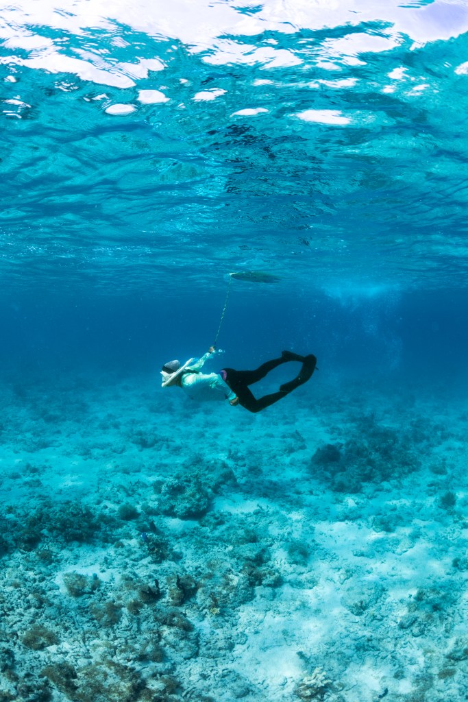 In this underwater image, a swimmer in a teal long sleeve shirt, black leggings and fins, and a snorkel swims through blue water halfway between the seafloor and the surface. Attached to her wrist is a thin cord, attached to a floatation device at the surface. The seafloor below is covered in rocks and pale sand.