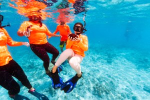 In this underwater image, a girl in a neon orange long-sleeve, white leggings, black fins, and black snorkel floats in the water, holding a round chunk of brown coral. Other students in the same orange shirts swim around her, against bright blue water in the background.
