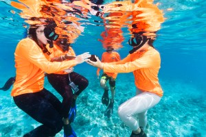In this underwater image, a girl in a neon orange long-sleeve, white leggings, black fins, and black snorkel floats in the water, holding a round chunk of brown coral out towards another student in the same orange shirt. Other students in the same orange shirts swim around her, against bright blue water in the background.