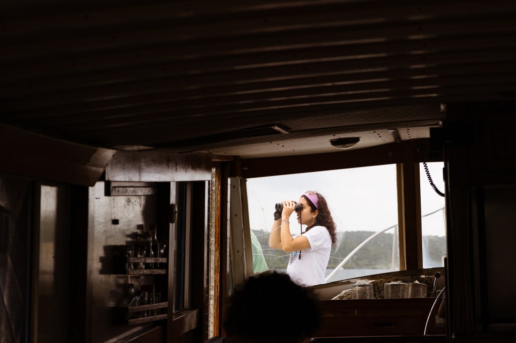 A shot taken inside of a boat with one window visible. Out the window, a student with brown hair and a white t-shirt looks through binoculars, pointed to the left of the frame. Everything inside the boat is dark brown wood.