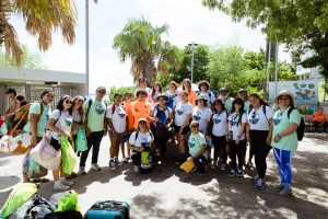 A group photo of students and instructors wearing white, teal, and orange shirts stand together in an outdoor area, with buildings and trees in the background.