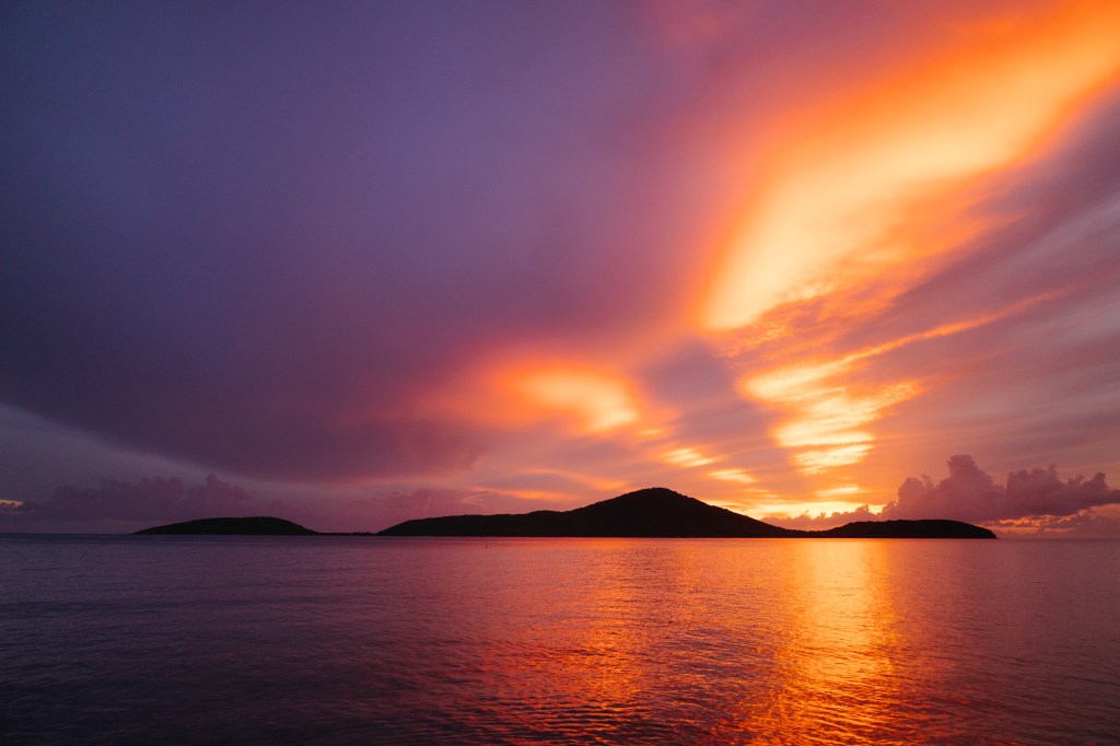 An image of a sunset with bright yellow and orange concentrated towards the right of the frame, fading to deep purple towards the left. The lower third of the image is water, reflecting the sky above, with the thin black silhouettes of a mountainous island on the horizon.