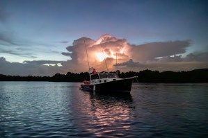 A small black and white boat floats in a bay during blue hour, with a lightning storm directly behind the boat.