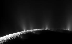 A black and white image of Enceladus' south pole. Several plumes of water ice spray up from the curved surface; they are visible as misty white spikes. Most of the south pole is dark, but a small sliver is lit up at bottom left.