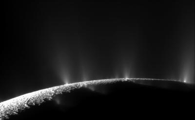 A black and white image of Enceladus' south pole. Several plumes of water ice spray up from the curved surface; they are visible as misty white spikes. Most of the south pole is dark, but a small sliver is lit up at bottom left.