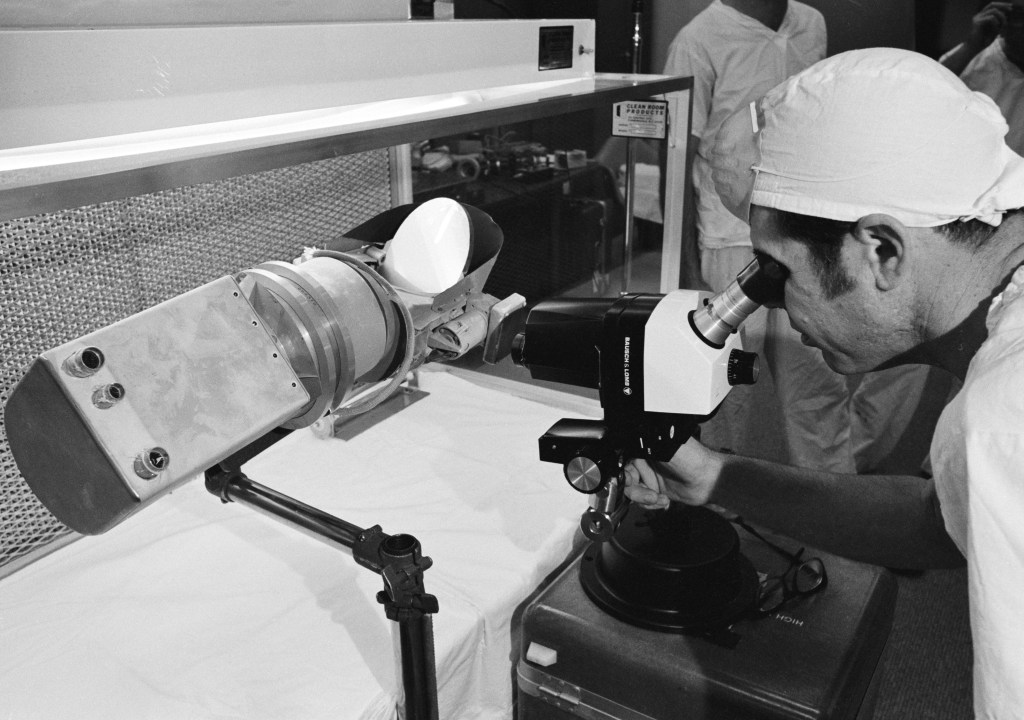 View of scientist looking at a Surveyor III components with a microscope in a Lunar Receiving Laboratory sterile vacuum cabinet.