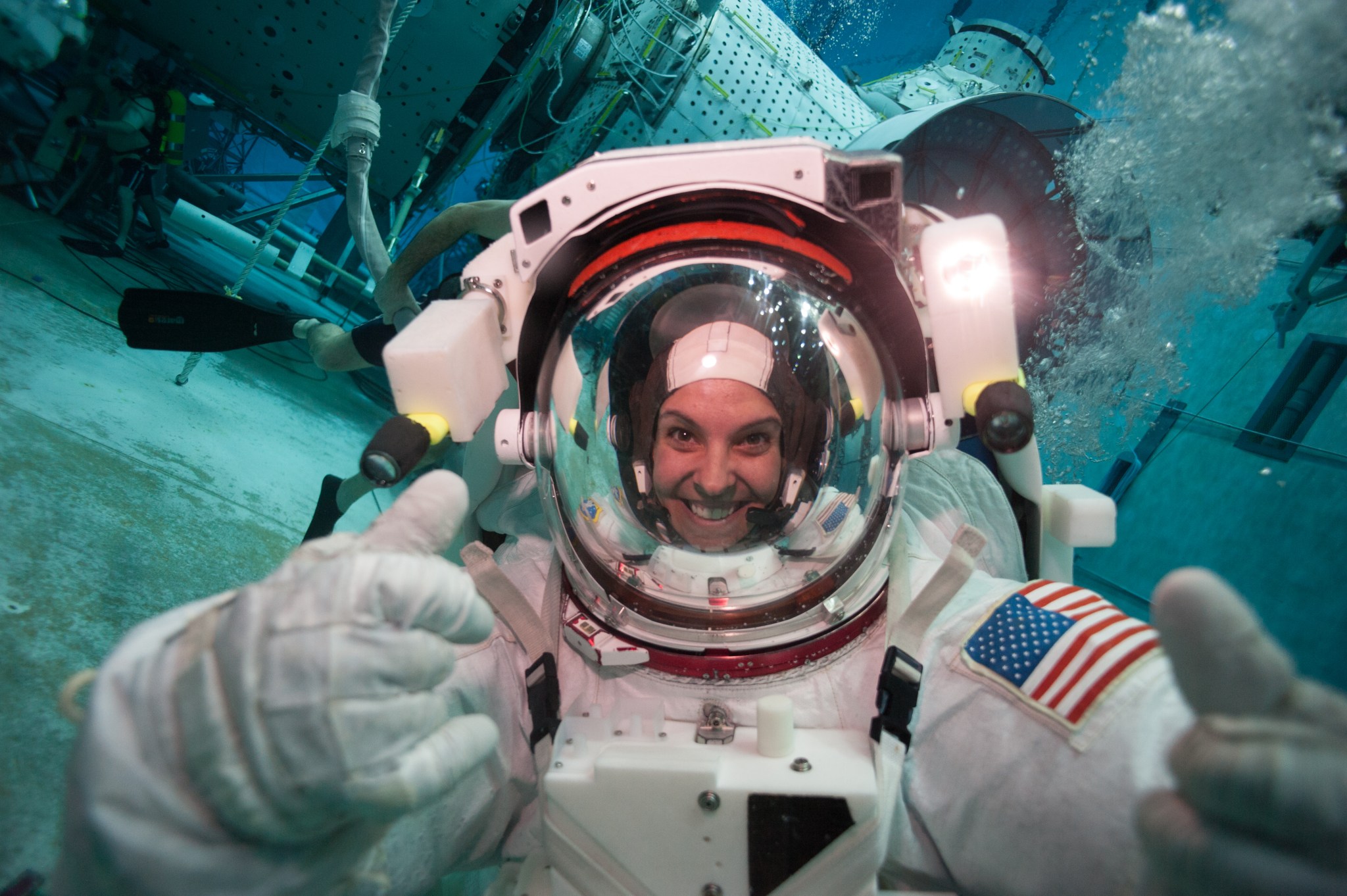 A woman smiles and gives a thumbs up underwater in a spacesuit.