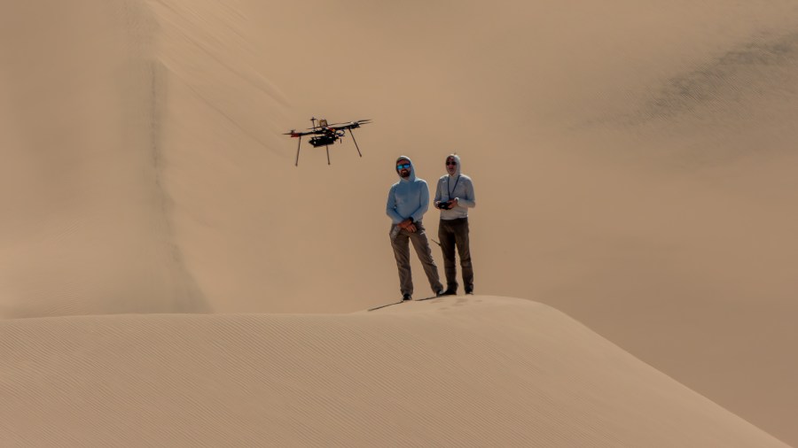 Two people stand side by side on a sandy hill, or dune. The person on the left is standing in a blue top, while the person on the right in a gray top is holding a controller. Above and to the left of their heads is a rotorcraft flying above the dune. The background of this image is more sandy dunes.