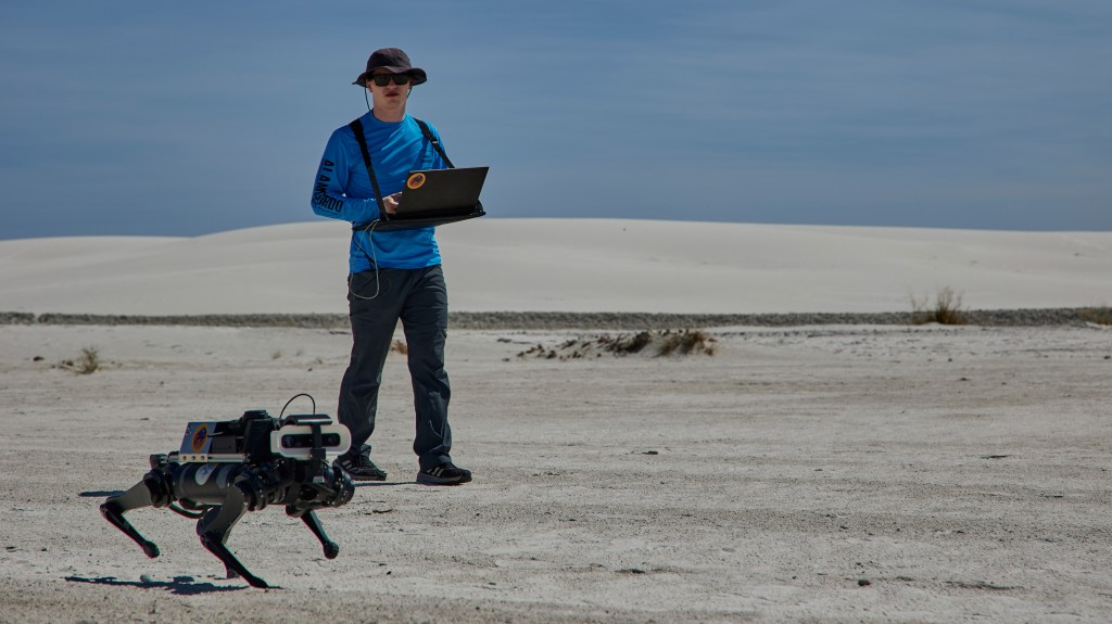 One person in a blue shirt and a bucket hat walks on a flat, white sandy ground and there is a blue sky behind them. They are holding a laptop on a harness around their neck while in the foreground, a small, dog-like robot walks.
