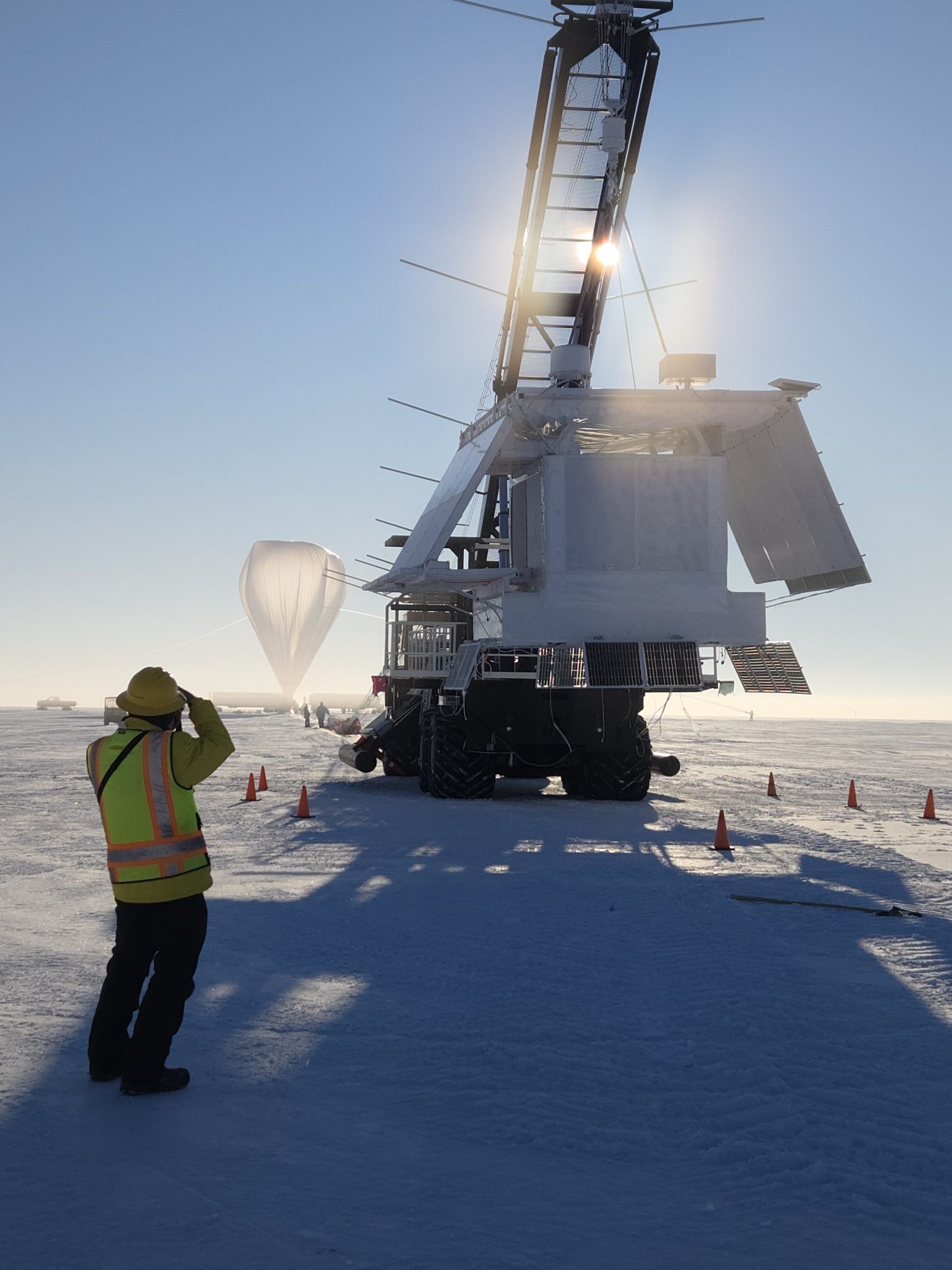 A man in a yellow safety vest looks at a scientific balloon in the distance. In the foreground is the GAPS payload hanging from a crane preparing for launch.