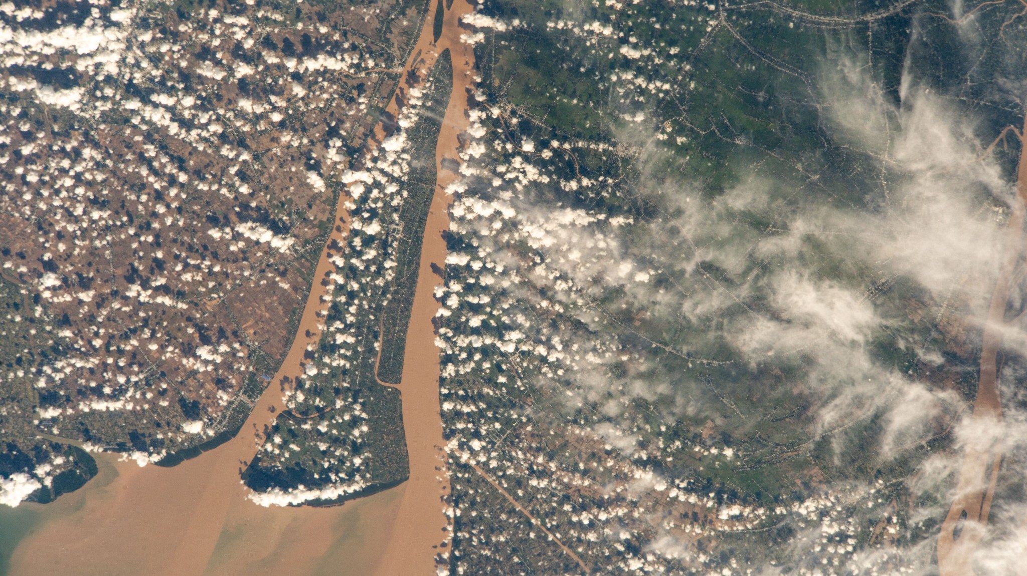 A view of a brown river and an islet from the International Space Station. The land visible on the right is mostly dark green, while the land on the left is brown with green edges along the river. Clumps of smaller white clouds dot the photo, almost like lines of dots. Wispier clouds are visible along the right side of the image.