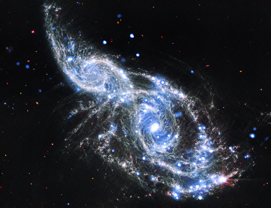 A pair of colliding spiral galaxies. Here, both spirals are shown face on, with the smaller of the two galaxies, IC 2163, at the upper left of the larger galaxy, NGC 2207, which dominates the center and lower right of the image. Both galaxies have long, spiraling, silver blue arms, dotted with specs of blue and red. Toward our upper left, the curving arms overlap, and bend toward their neighbors' core. James Webb mid-infrared image (white, gray, and red) and adds the X-ray view from Chandra (blue).