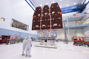 Two people in white jumpsuits look up at a large telescope with three black and orange panels facing them. They are in a large white cleanroom with equipment and scaffolding.
