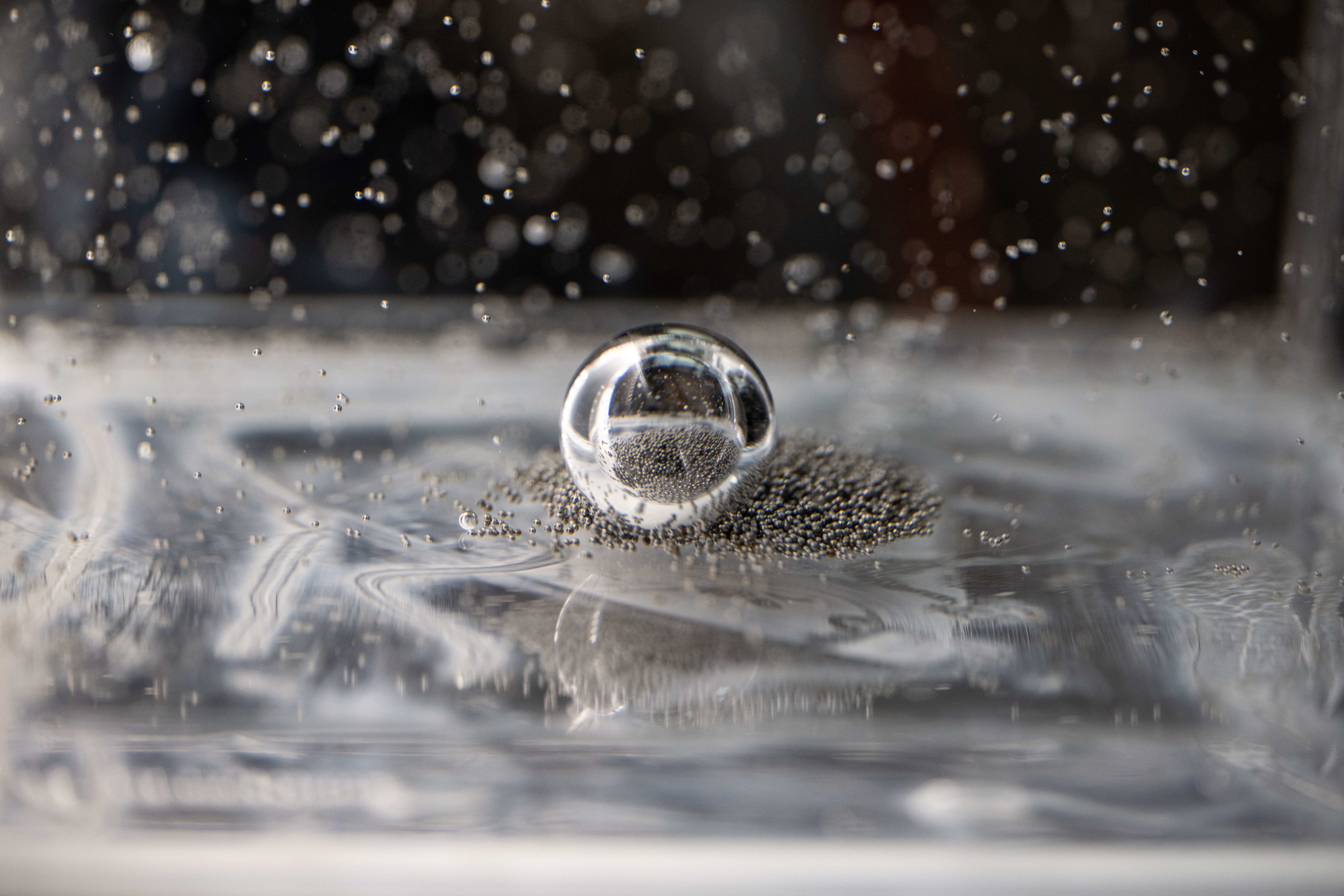 Tiny silver ball bearings crowd around a larger bearing in a clear, viscous fluid. Some of the smaller ball bearings are suspended in the fluid.