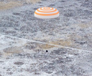 An orange and white striped parachute attached to a black spacecraft. The spacecraft is very small compared to the parachute. It is landing on ground that has white frost on it.