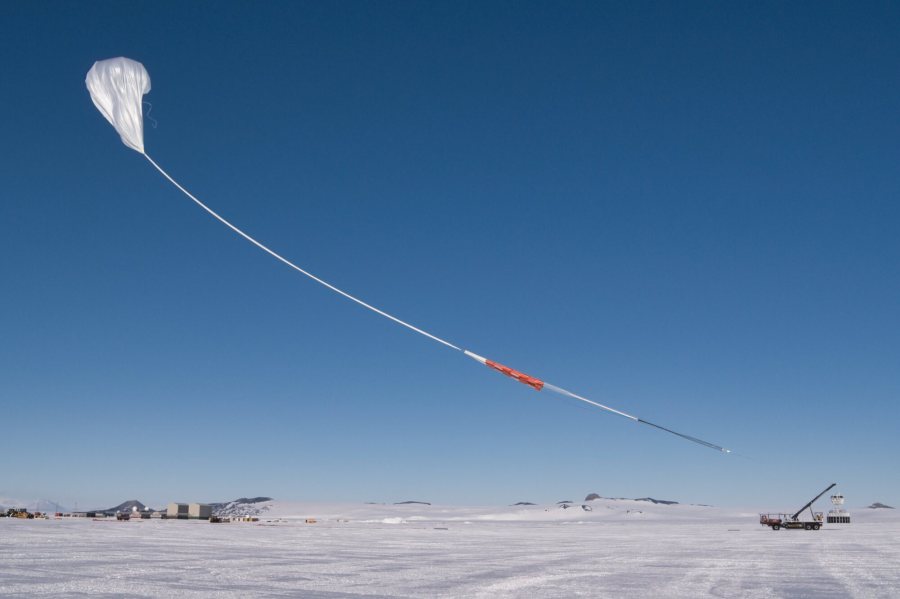 A white scientific balloon rises into the bright blue sky above snowy Antarctica. The sky and the balloon take up most of the image, with the ground only being a small white stripe at the bottom of the photo. The balloon has a long white "tail" with orange and black portions.