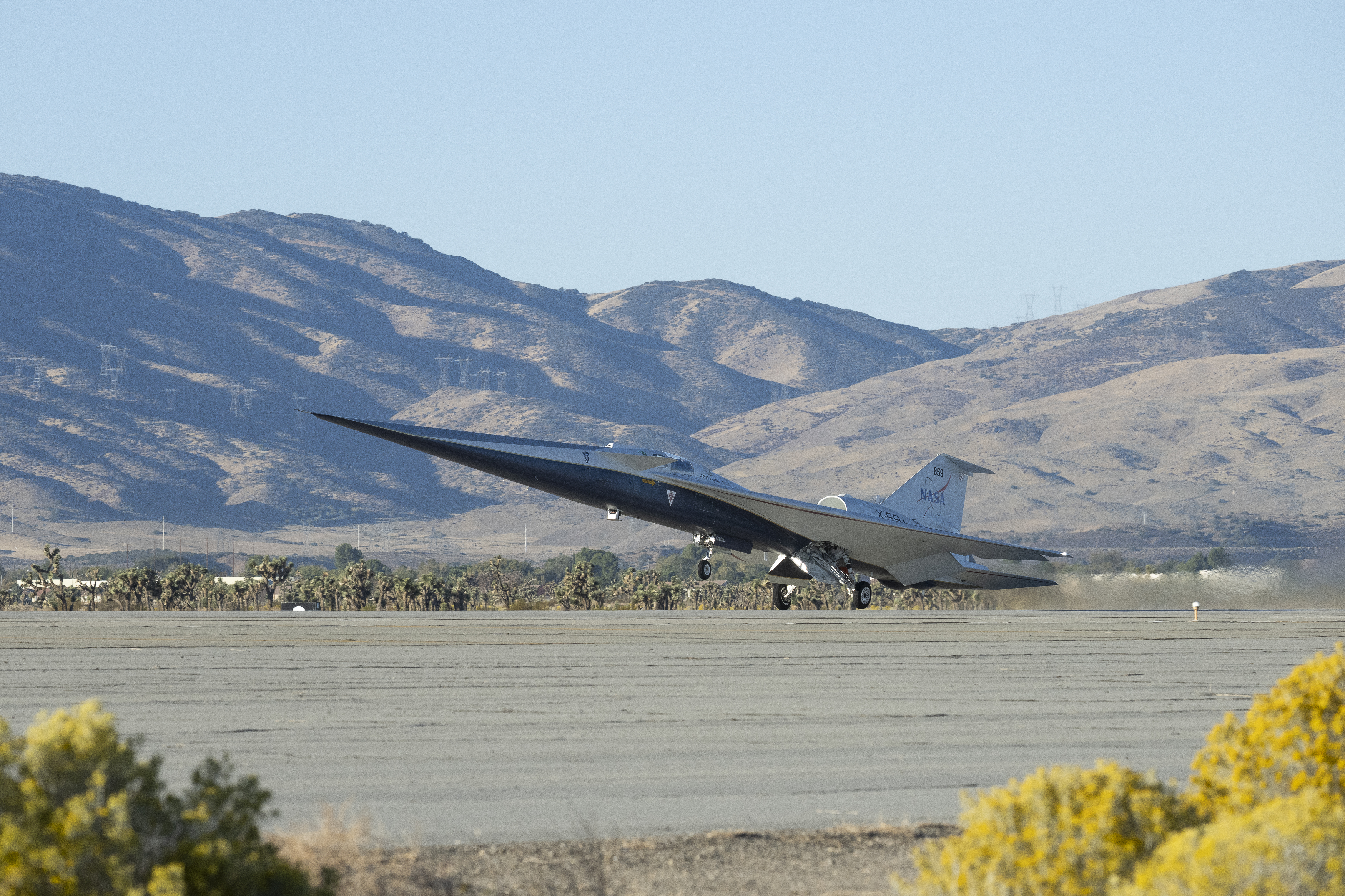 NASA’s X-59 quiet supersonic research aircraft lifts off from the runway at U.S. Air Force Plant 42 in Palmdale, California. Its wheels are just slightly off the runway. Heat waves are visible off the back of the aircraft as it ascends under a clear sky. Rows of Joshua trees stretch toward the mountains in the background.