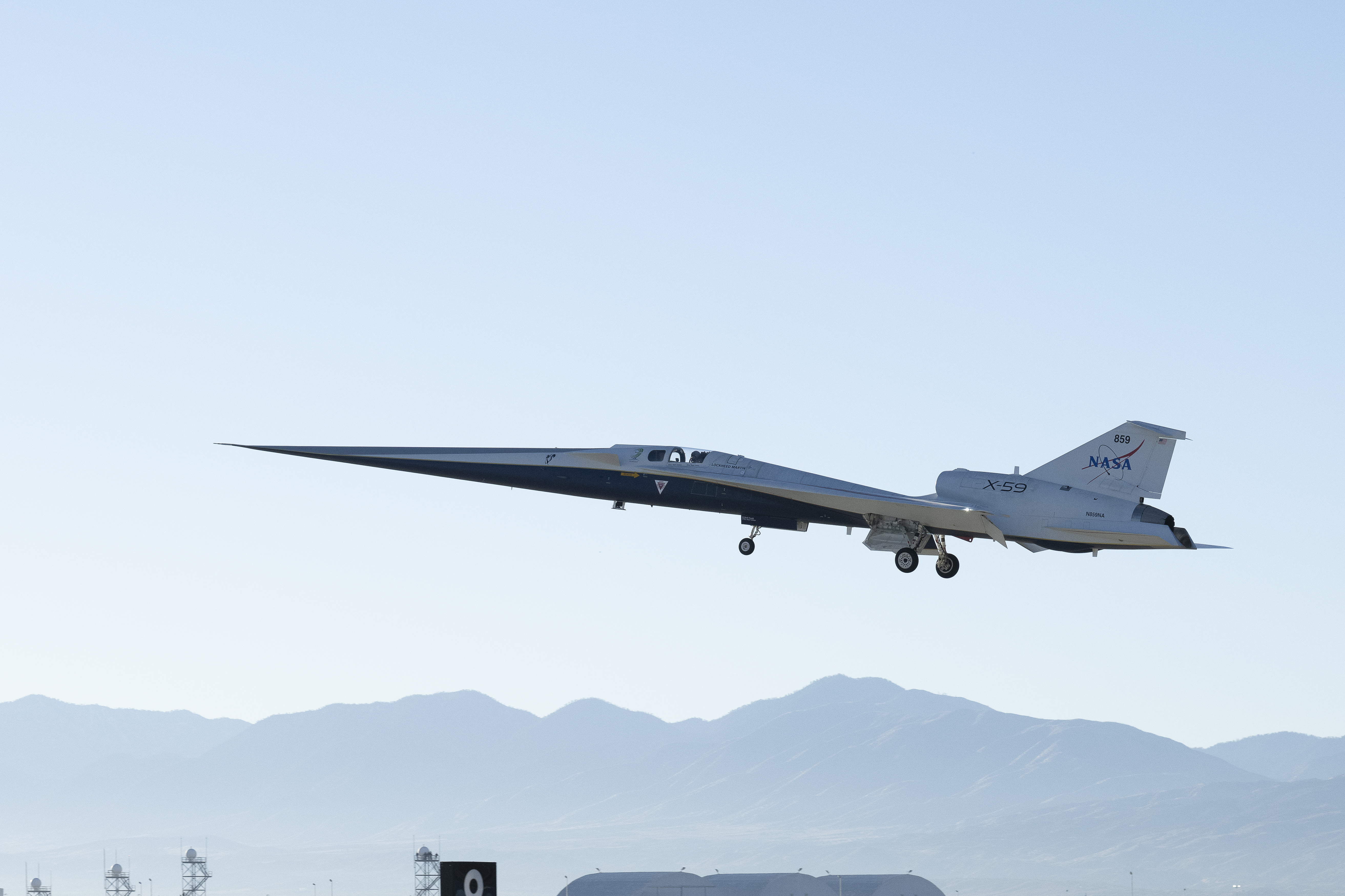 NASA’s X-59 quiet supersonic research aircraft lifts off from the runway at U.S. Air Force Plant 42 in Palmdale, California. The nose is angled slightly upward, and the landing gear is down as it ascends under a blue sky. The tops of buildings are visible below, with mountains in the far background.