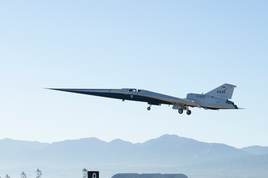 NASA’s X-59 quiet supersonic research aircraft lifts off from the runway at U.S. Air Force Plant 42 in Palmdale, California. The nose is angled slightly upward, and the landing gear is down as it ascends under a blue sky. The tops of buildings are visible below, with mountains in the far background.