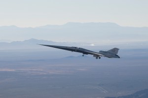 NASA’s X-59 flies above the Mojave Desert against a blue sky. The aircraft’s long nose is angled slightly upward to the left, with its landing gear down. Layers of mountains are visible in the background. The aircraft is backlit, appearing to float as it ferries to NASA’s Armstrong Flight Research Center in Edwards, California.