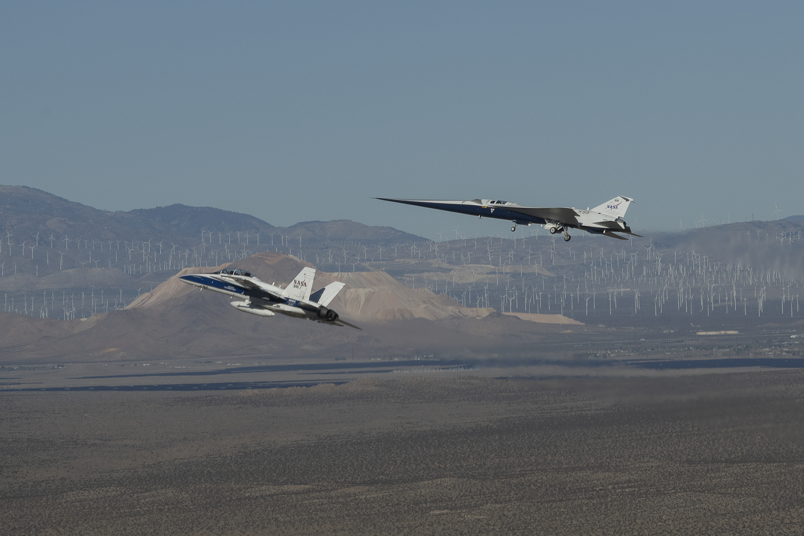 NASA’s X-59 flies above the Mojave Desert with a NASA F-15 off its left wing. The X-59’s landing gear is down and below it the desert landscape stretches toward rows of windmills in front of mountains.